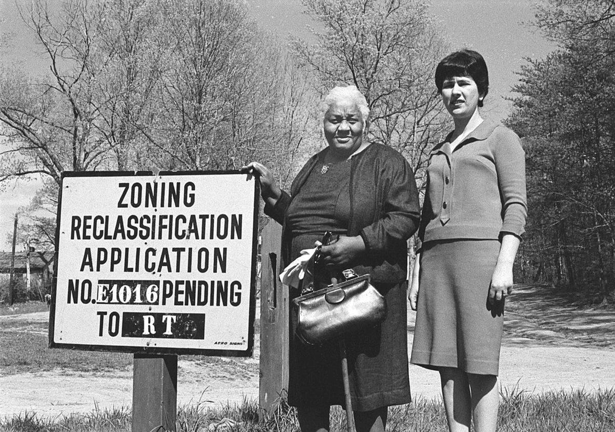 Black-and-white photograph of two women standing beside a roadside sign that reads “Zoning Reclassification Application No. E1016 Pending”
